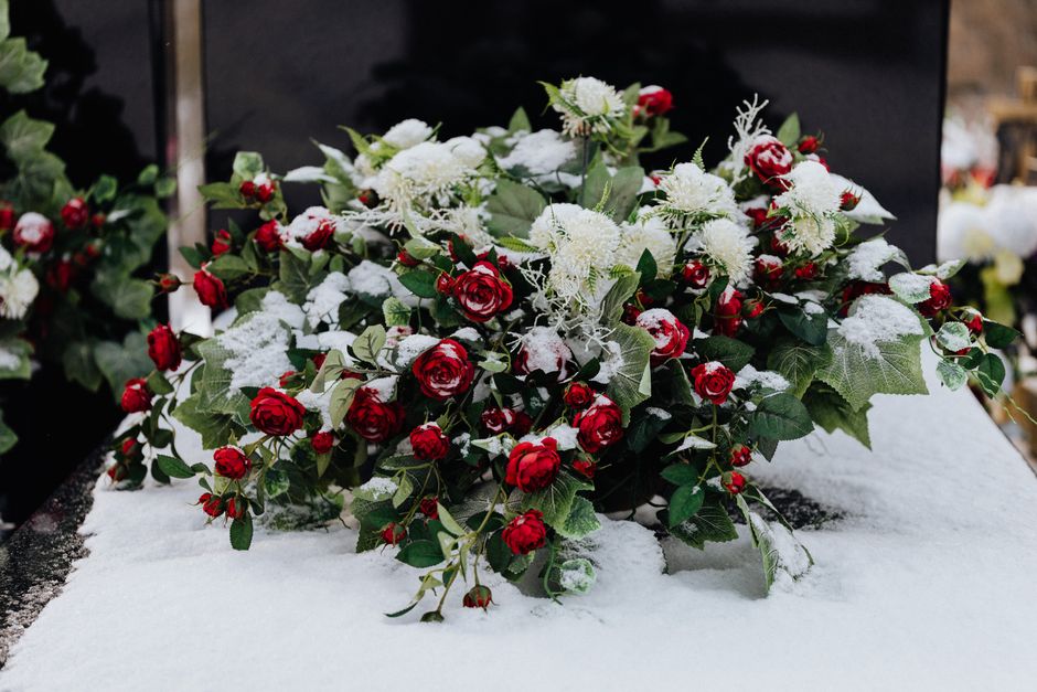 A floral arrangement of fresh red roses and white chrysanthemum flowers with green holly leaves, wrapped in clear cellophane and topped with a white satin ribbon, is laid on a snow-covered surface out