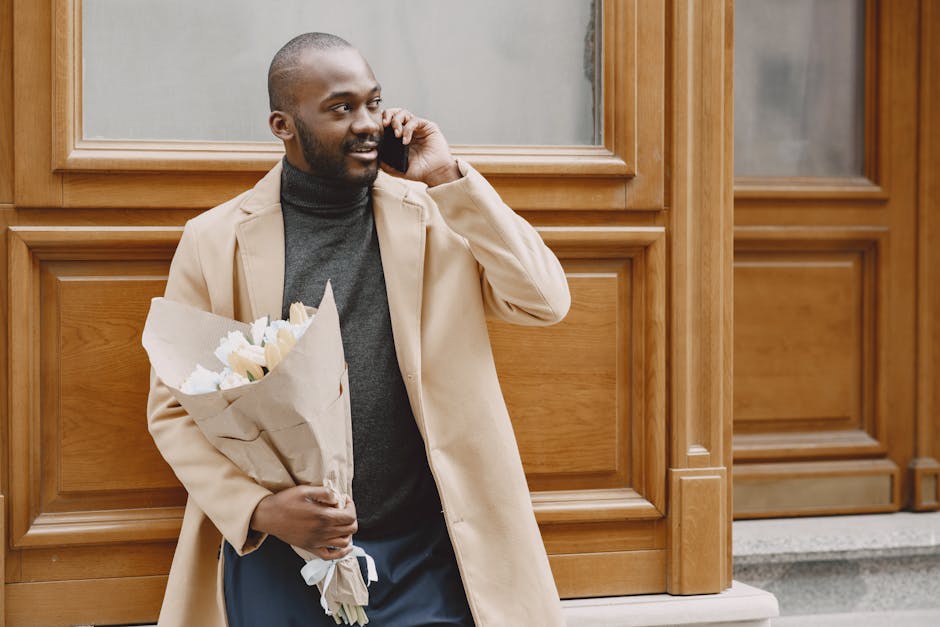 A man with a shaved head and a beard is standing outdoors in front of a wooden door with decorative panels, holding a bouquet of fresh white roses wrapped in brown paper. He is wearing a beige overcoa