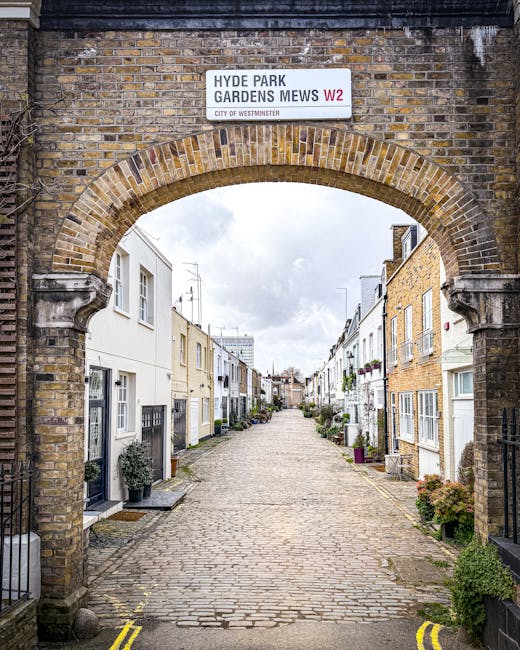 A charming narrow cobblestone street viewed through a decorative brick archway, with a white sign reading 'Hyde Park Gardens Mews W2' overhead. On either side of the street, there are row houses with 