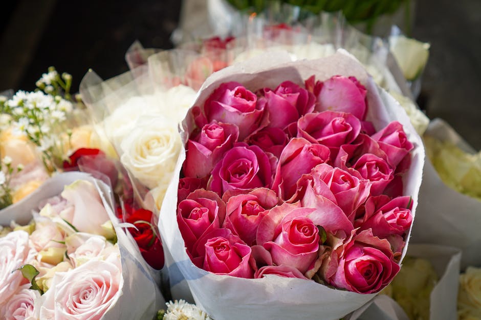 A close-up of a floral gift bouquet featuring a dense cluster of pink roses with delicate, velvety petals, arranged in a white paper wrapping. The bouquet rests on a surface alongside other flower arr