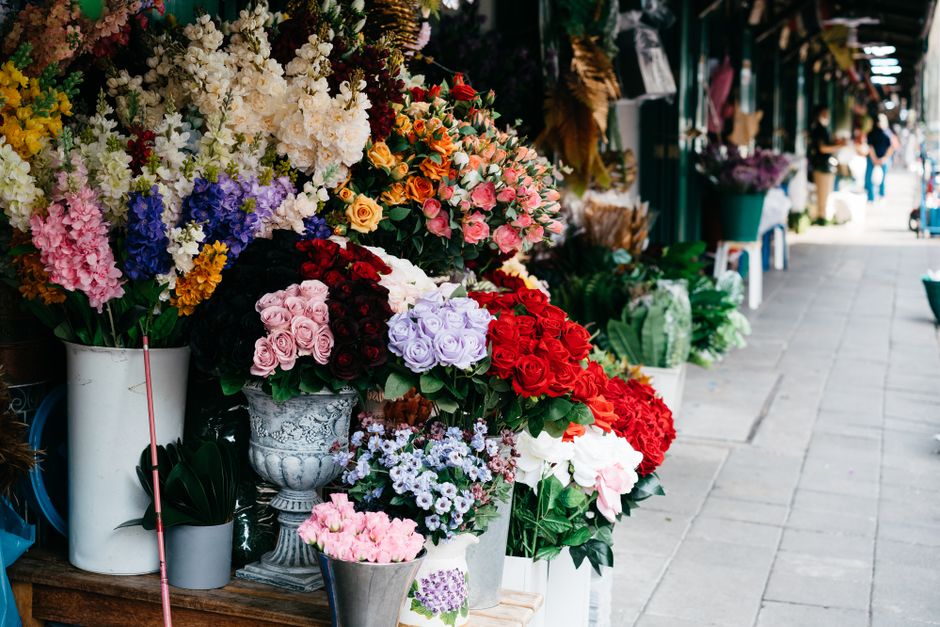 A vibrant outdoor flower shop display featuring an assortment of fresh floral bouquets arranged in white ceramic and metal vases. Prominent flowers include pink roses, deep red roses, lavender-colored